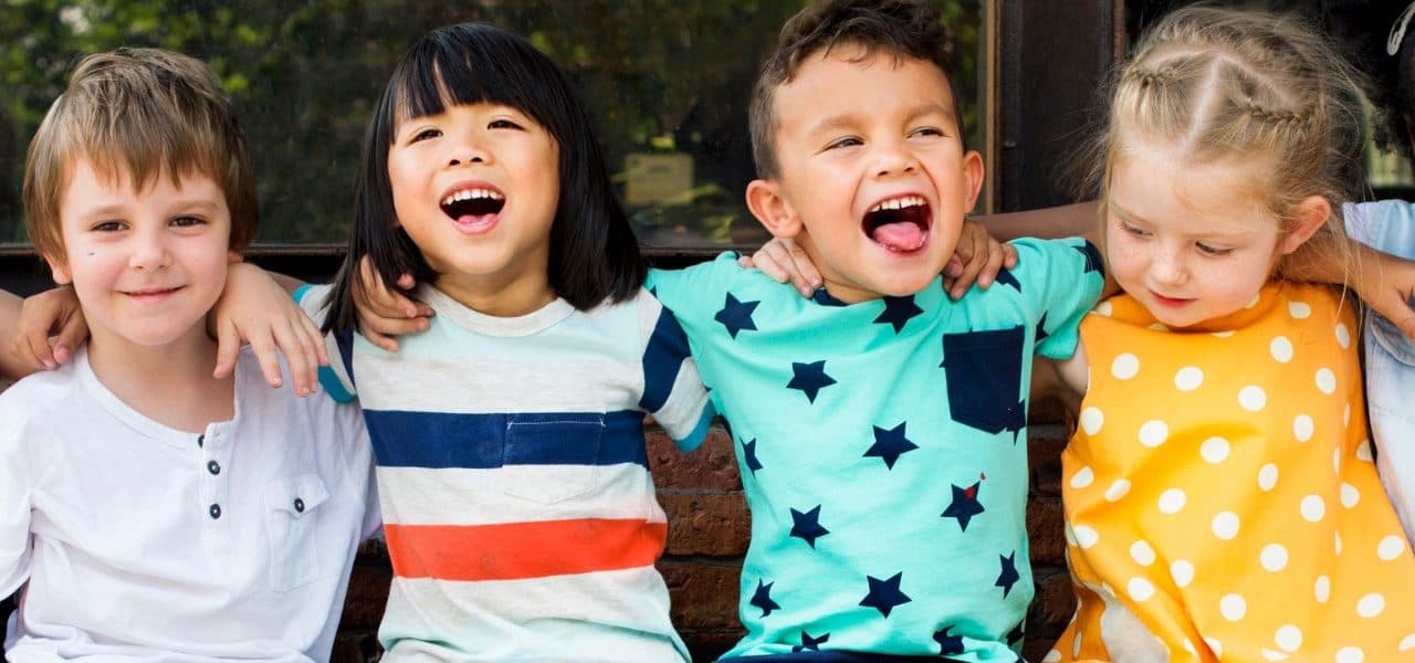 Four young children sit side by side with their arms around each other, laughing.